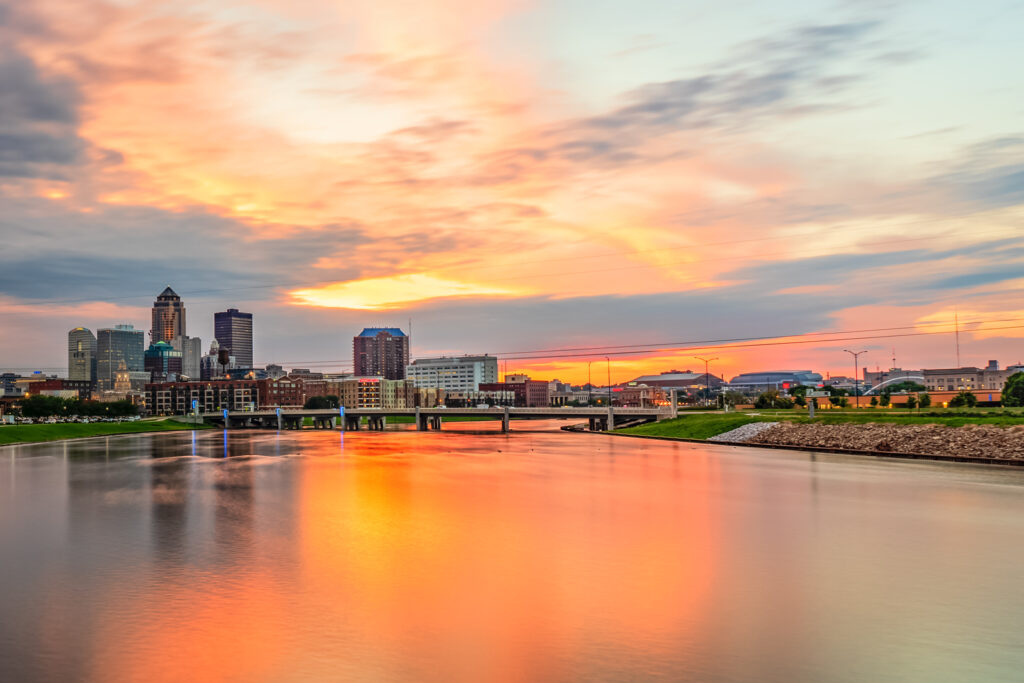  breathtaking photo of the downtown Des Moines skyline at sunset, with the city's modern skyscrapers silhouetted against a vibrant orange and pink sky, reflecting on the calm river below.