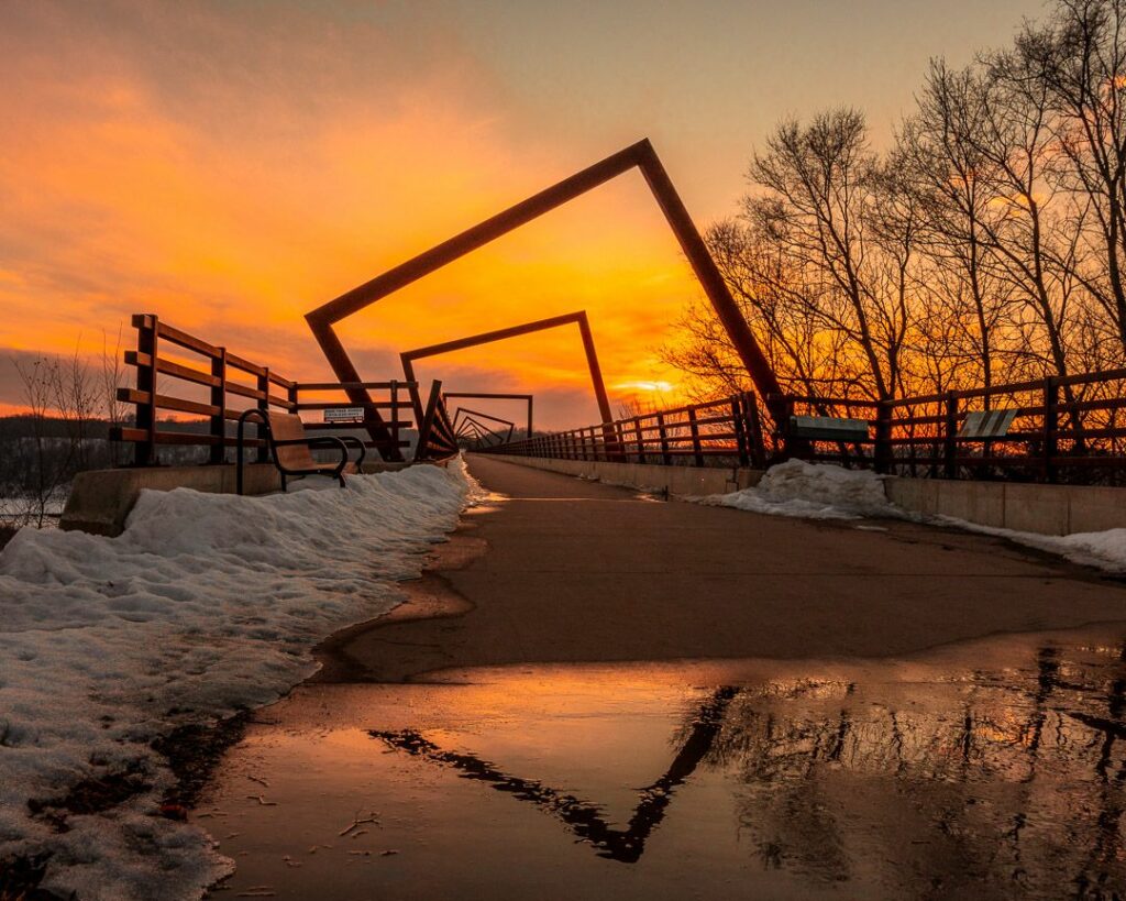 Stunning sunset view of the High Trestle Trail Bridge, featuring a reflection of its unique upper frame in a puddle of water on the bridge, creating a mesmerizing and unforgettable visual experience for visitors.