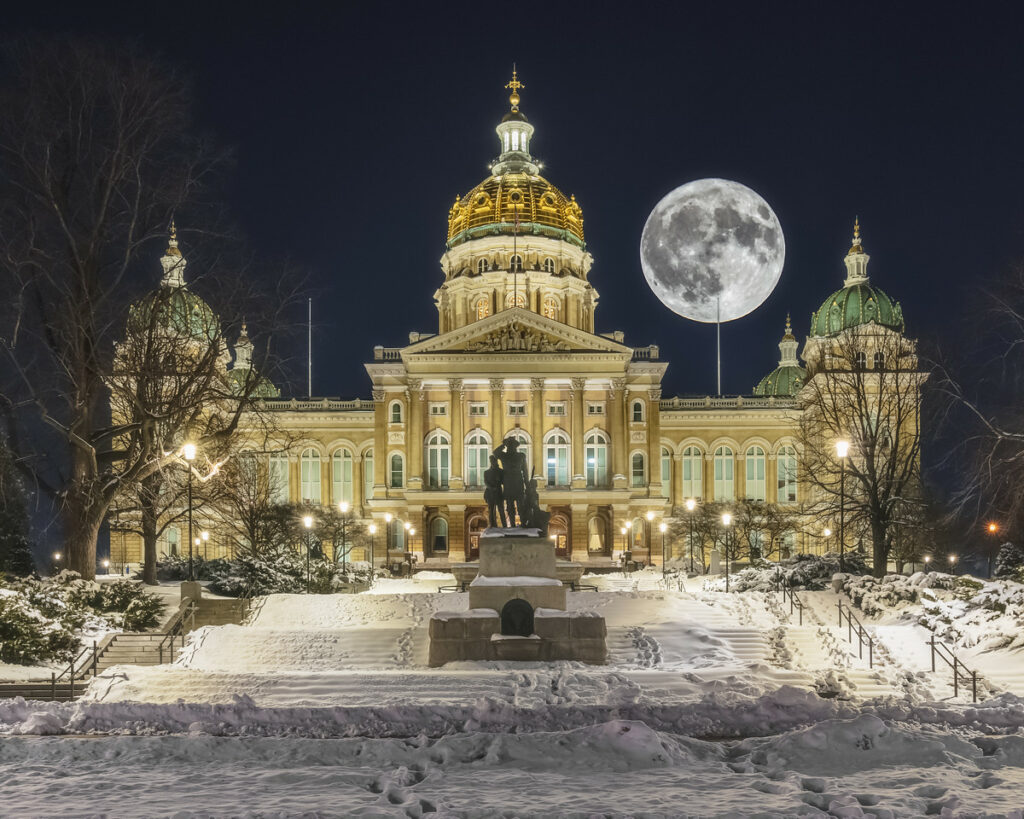 A stunning view of the Iowa State Capitol building, showcasing its golden dome and classical architecture, and a giant full moon. The ground is covered in snow.