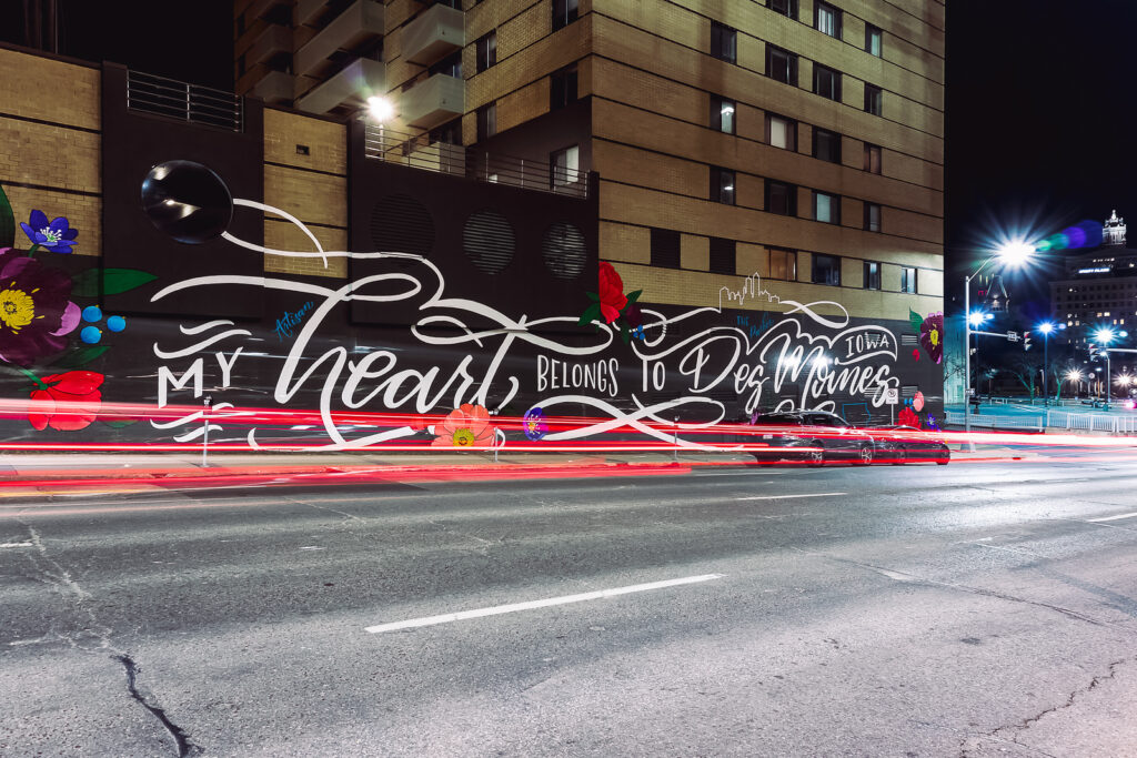 A dynamic long exposure shot of a building in downtown Des Moines featuring a vibrant mural that reads 'My Heart Belongs to Des Moines,' with streaks of red car taillights streaming down the street in front of the mural, symbolizing the city's energy and movement."