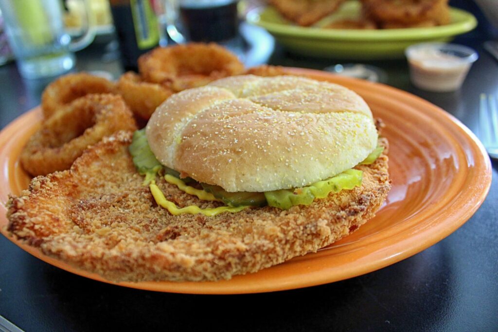 A photo of a gigantic pork tenderloin sandwhich with mustard and pickles on a sesame seed bun, served with onion rings.